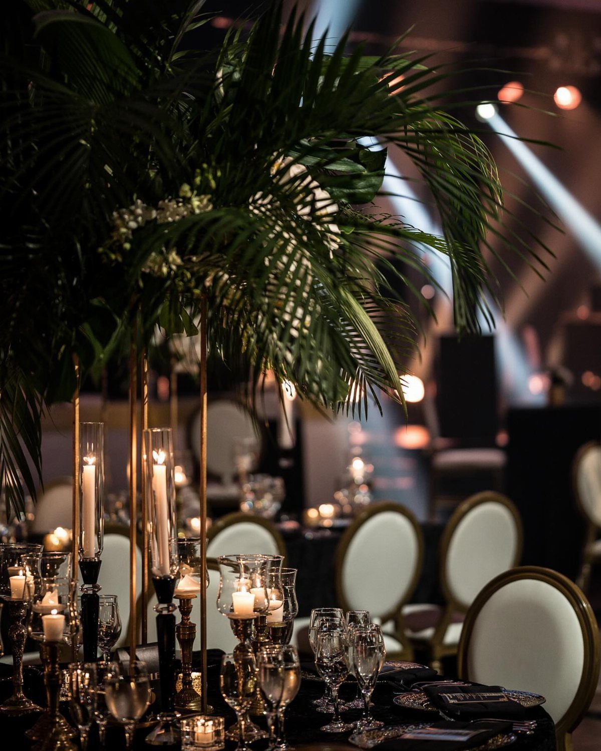 16 Marriage Theme Ideas for 2026: From Beach Ceremonies to Pearl Aesthetics 8 Tall gold centerpieces with palm leaves and white candles set on a dark wedding reception table.