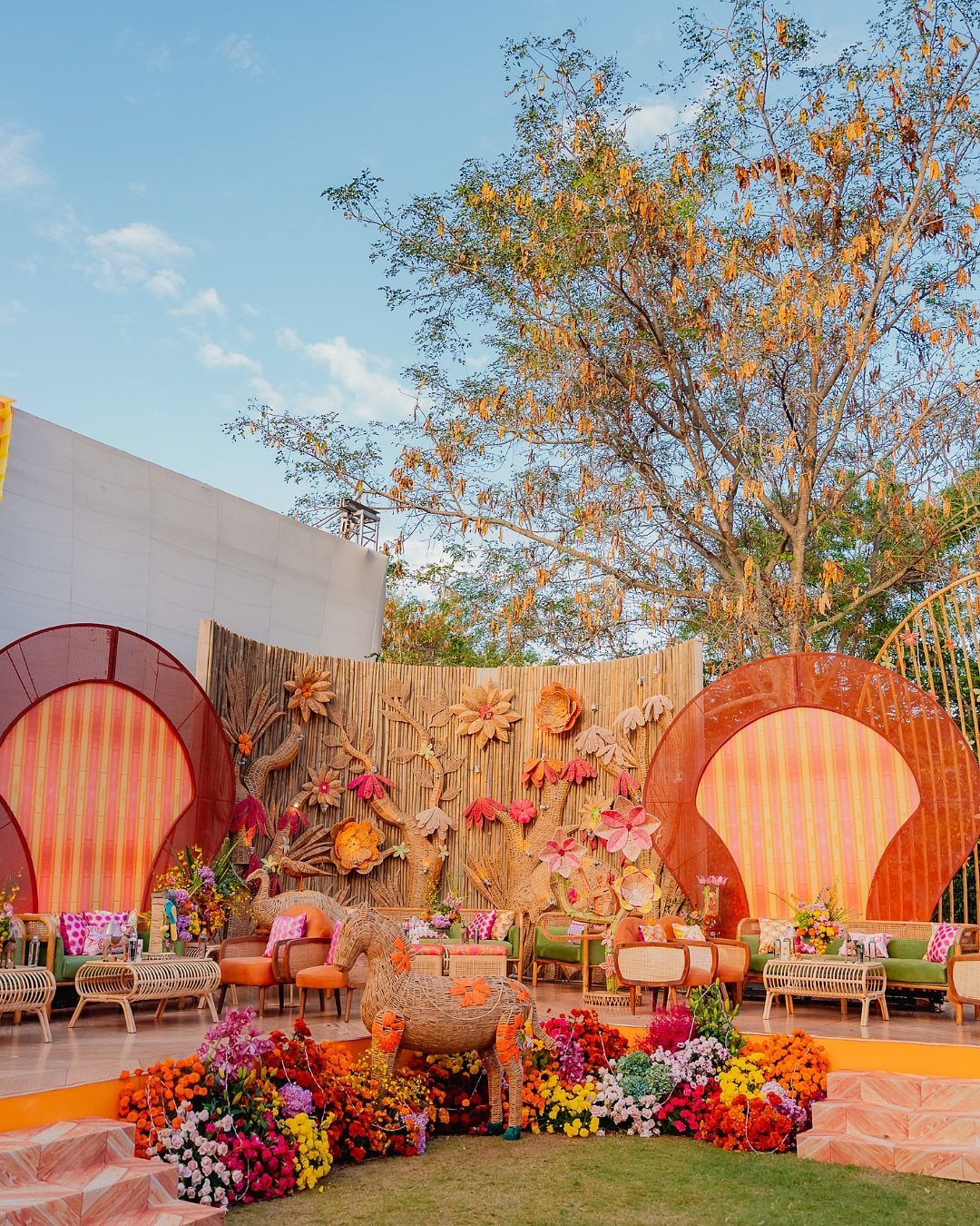 16 Marriage Theme Ideas for 2026: From Beach Ceremonies to Pearl Aesthetics 3 Colorful outdoor event decor with orange arches, bamboo walls, and vibrant floral arrangements on grass.