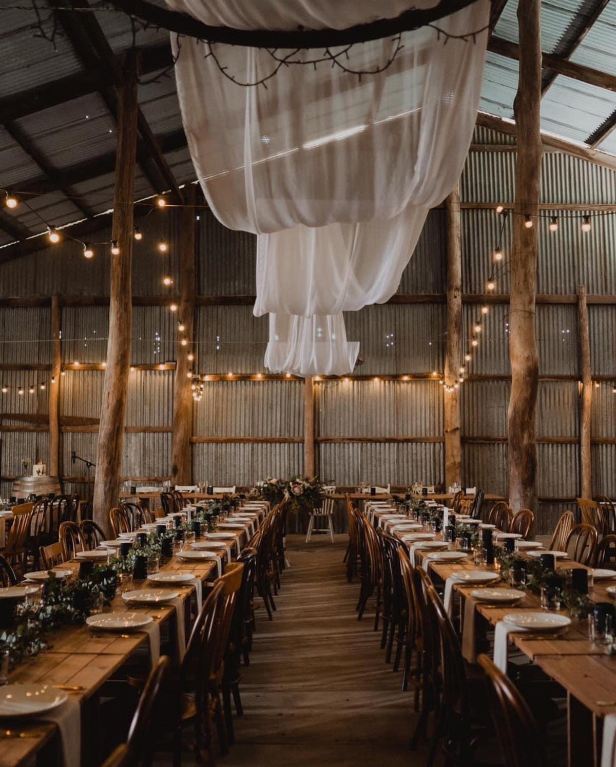16 Marriage Theme Ideas for 2026: From Beach Ceremonies to Pearl Aesthetics 1 Long wooden banquet tables set for a wedding inside a rustic barn with twinkling lights.