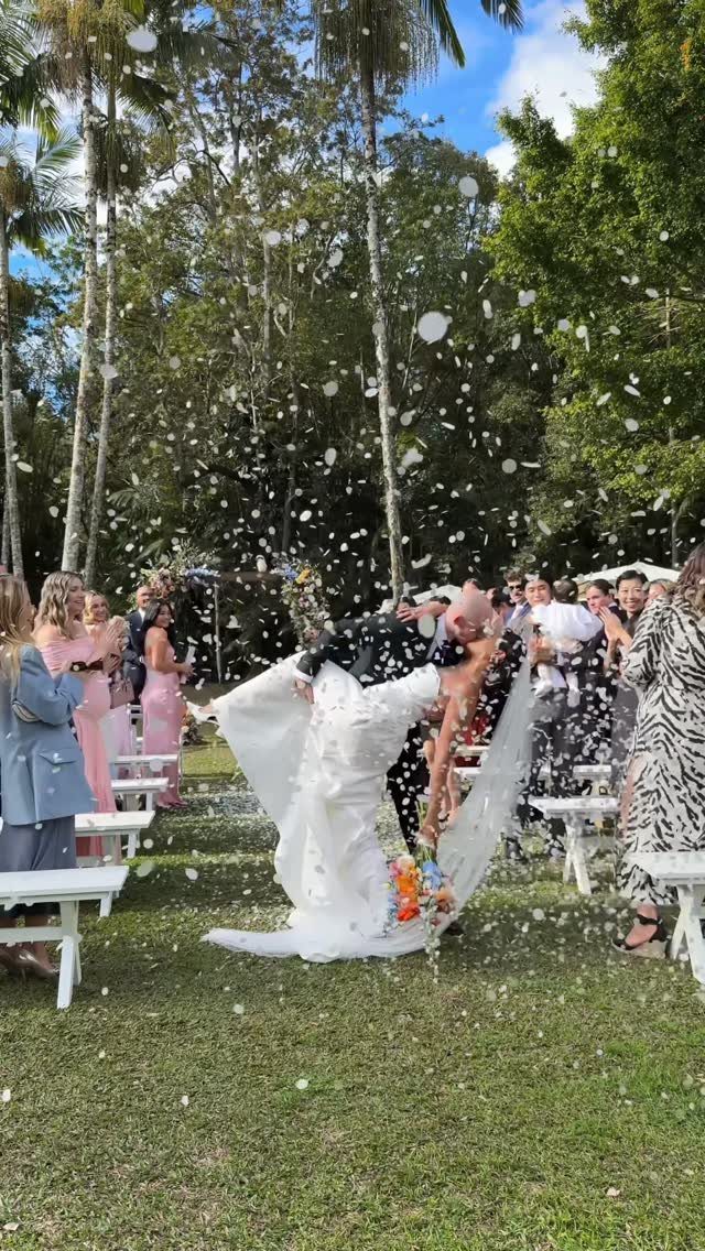 A bride and groom kissing outdoors amidst falling white flower petals and wedding guests.