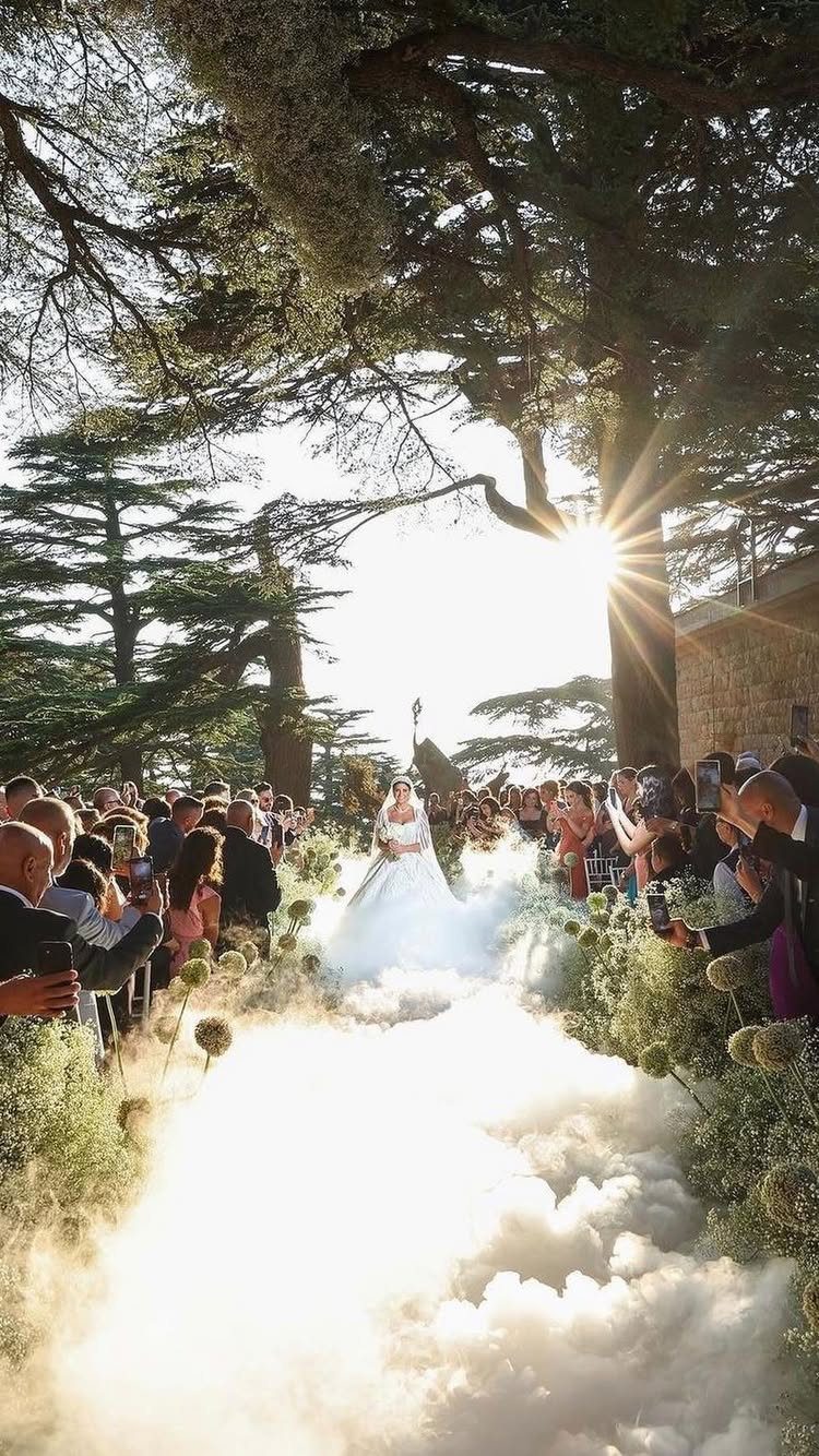 A bride walks down a white, cloud-like aisle lined by guests under large trees.