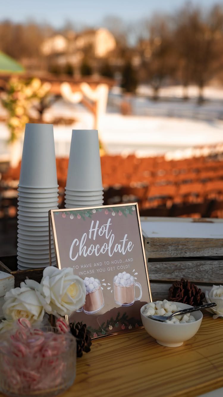 White paper cups and a hot chocolate sign sit on a wooden table with marshmallows and roses.