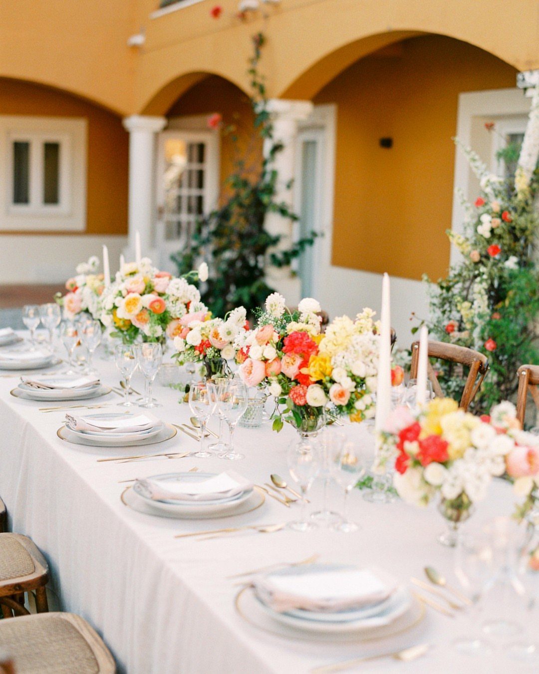 A long outdoor dining table decorated with colorful summer floral arrangements and white candles.