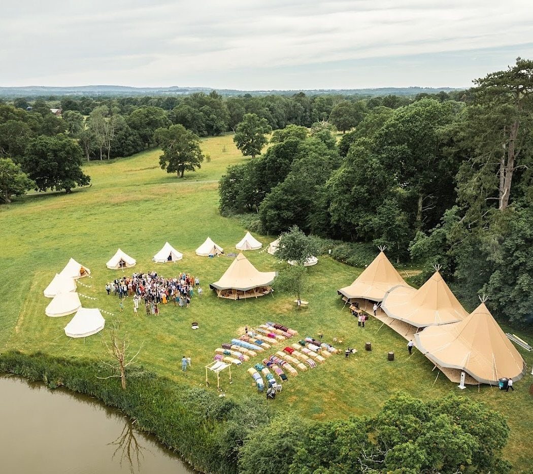 Aerial view of tan bell tents and people gathered on a green field by a river.