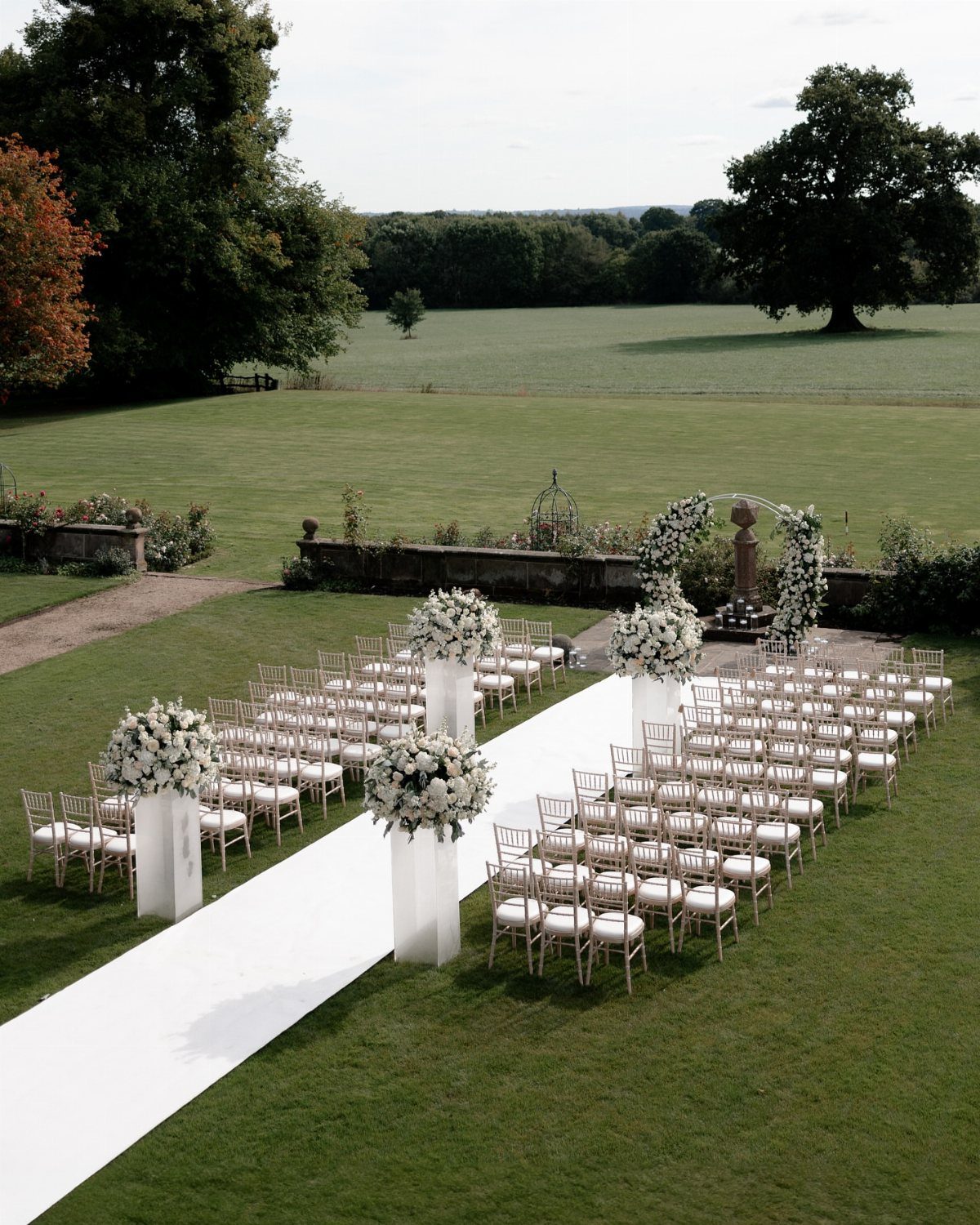 White wedding chairs and floral pedestals line a white aisle runner on a green lawn.