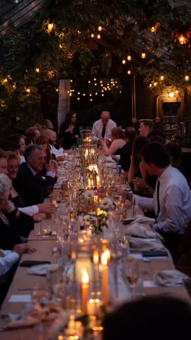 Guests seated at a long candlelit dinner table under a canopy of greenery and fairy lights.