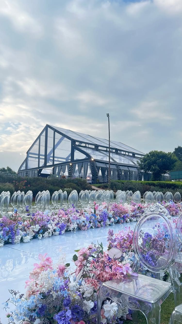 Transparent chairs and colorful floral arrangements line a white aisle runner near a large glass greenhouse.
