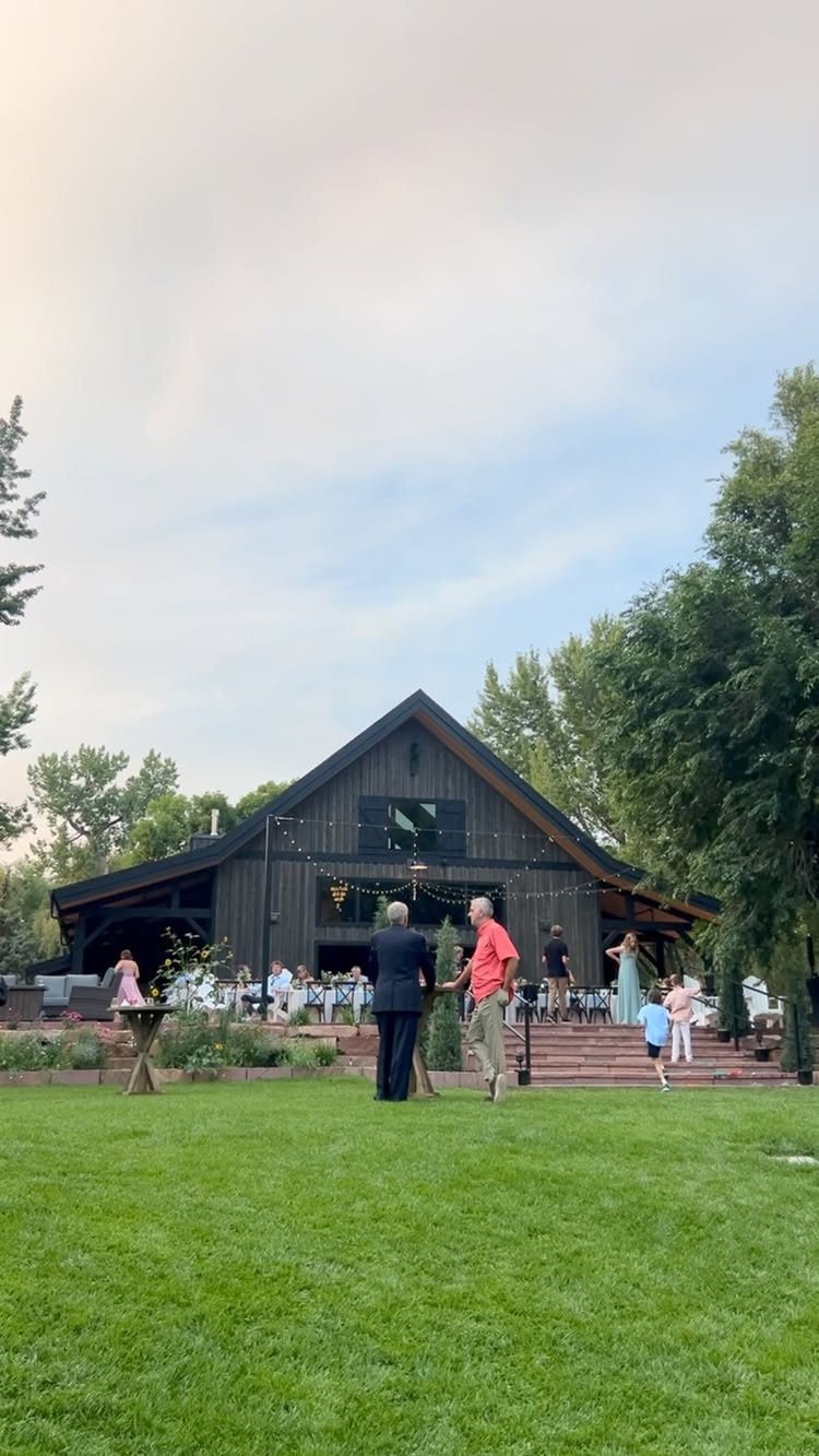A dark wooden barn decorated with string lights, surrounded by green grass and trees.