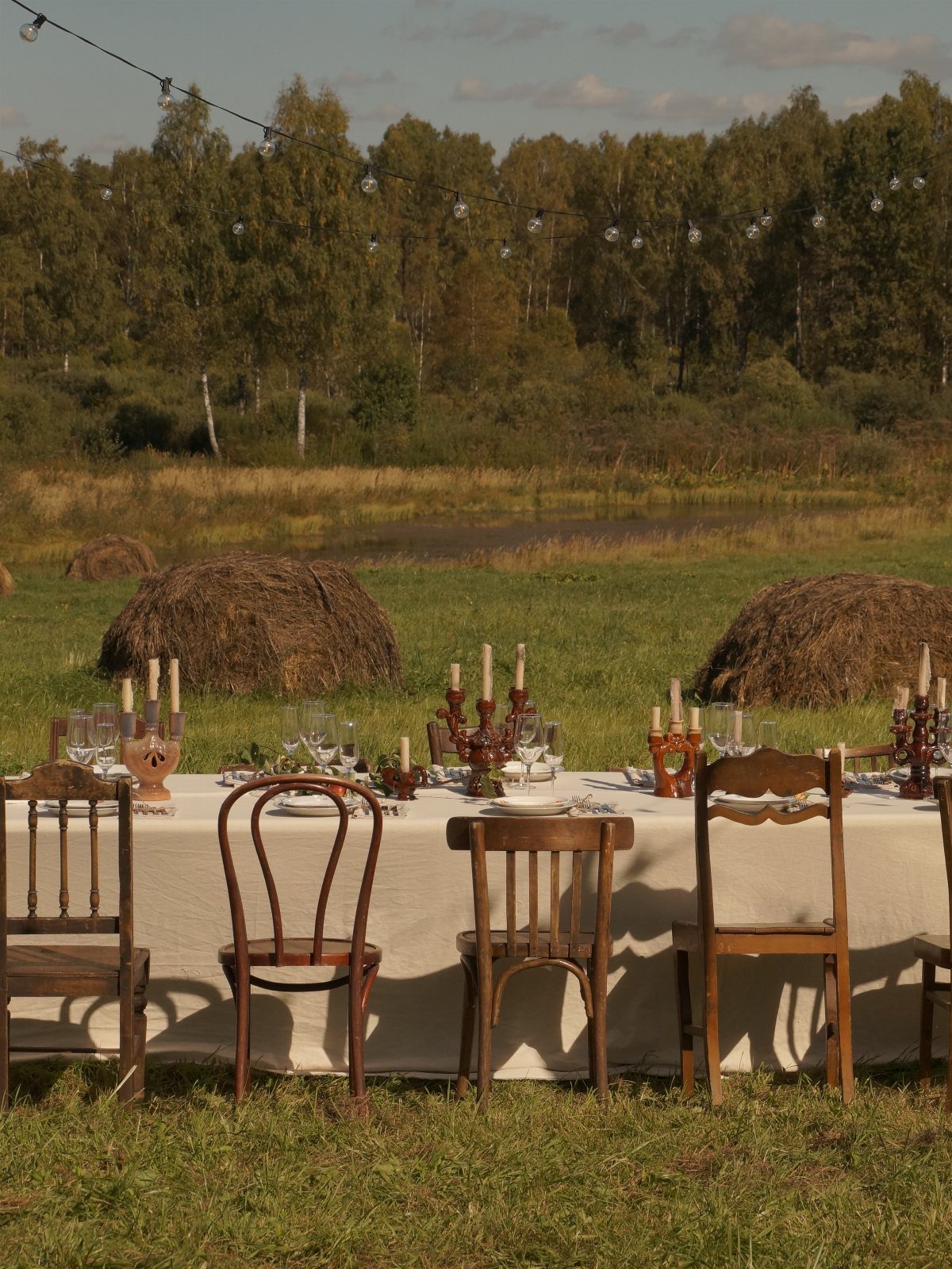 A long dining table set outdoors with wooden chairs and hay bales in a field.