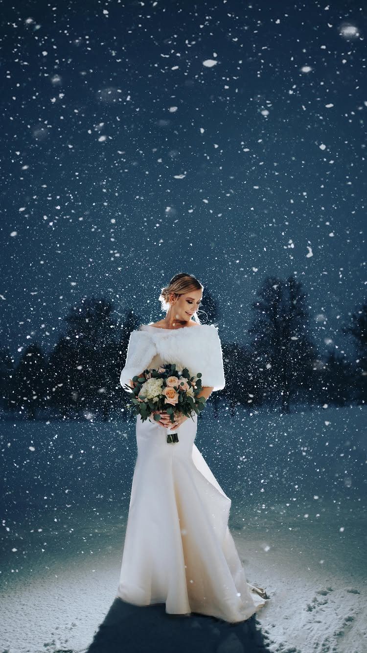A bride in a white gown holding a bouquet amidst falling snow at night.
