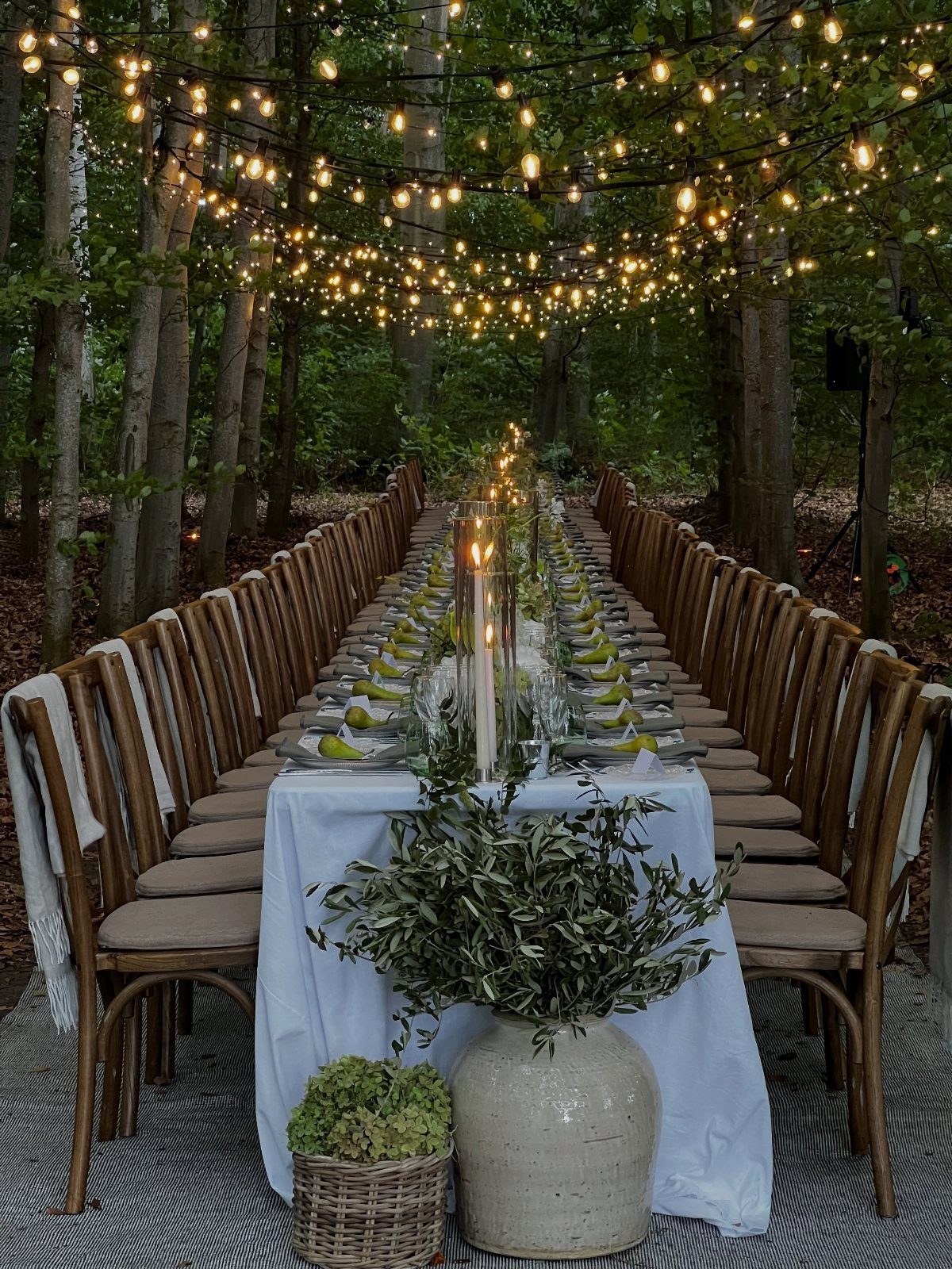 A long wedding dining table set with white linens and greenery under hanging string lights in a forest.