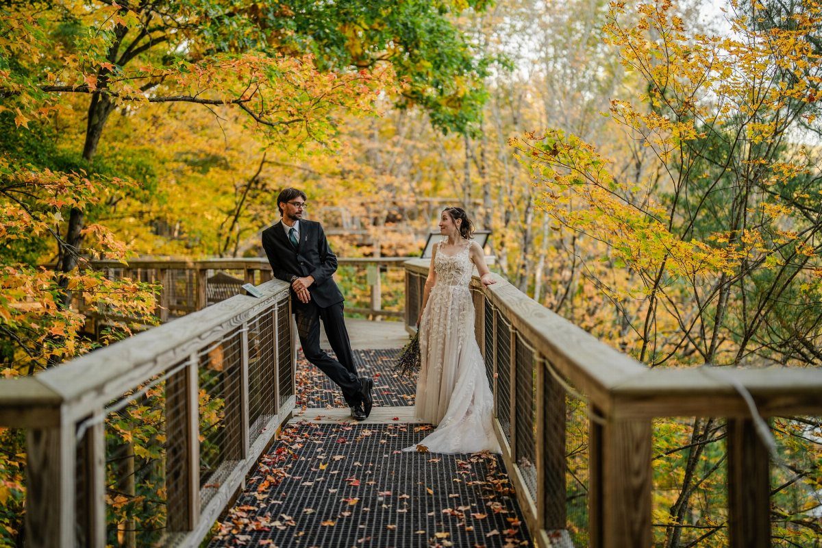 A bride and groom posing on a wooden forest walkway surrounded by autumn leaves.