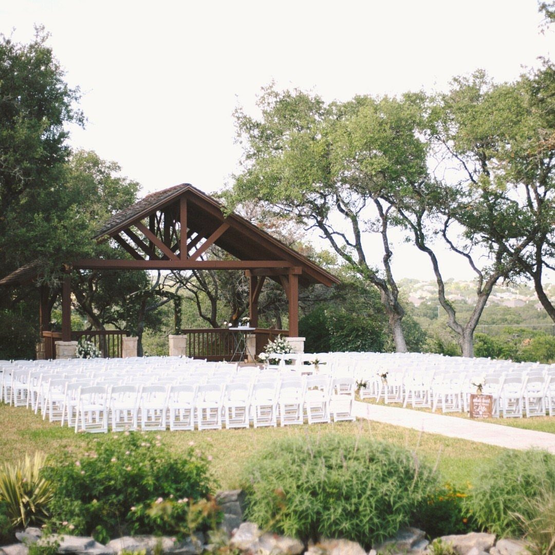 White folding chairs arranged in rows facing a wooden pavilion in a wooded outdoor setting.