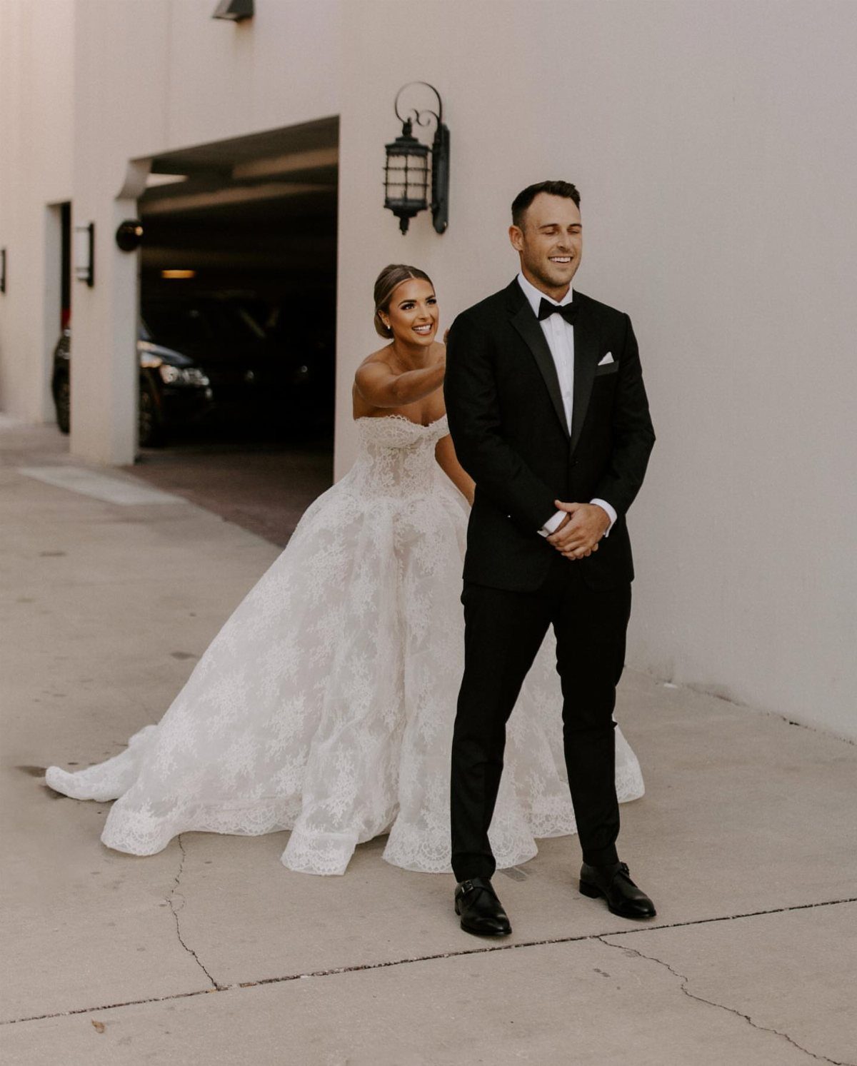 19 Photosession Ideas for Your Wedding Ceremony 2 A groom in a black tuxedo smiles while a bride in a white lace gown approaches him.