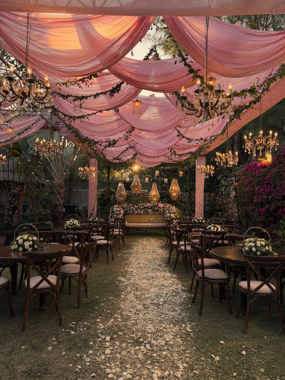 8 Pink Wedding Color Ideas for Every Venue and Season 8 Pink fabric drapes and chandeliers hang over an outdoor wedding reception with wooden tables and chairs.