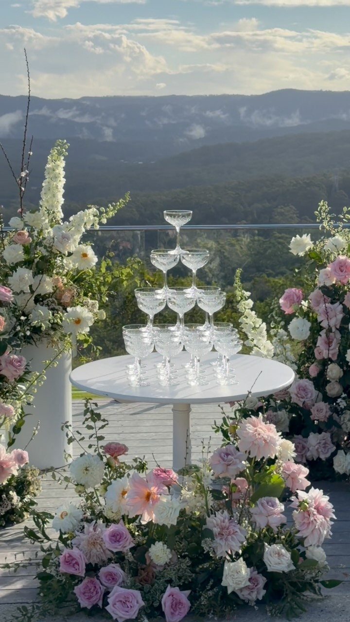 12 Romance Wedding Theme Ideas That Actually Feel Personal 10 Champagne glasses stacked in a tower on a white table surrounded by pink and white flowers.