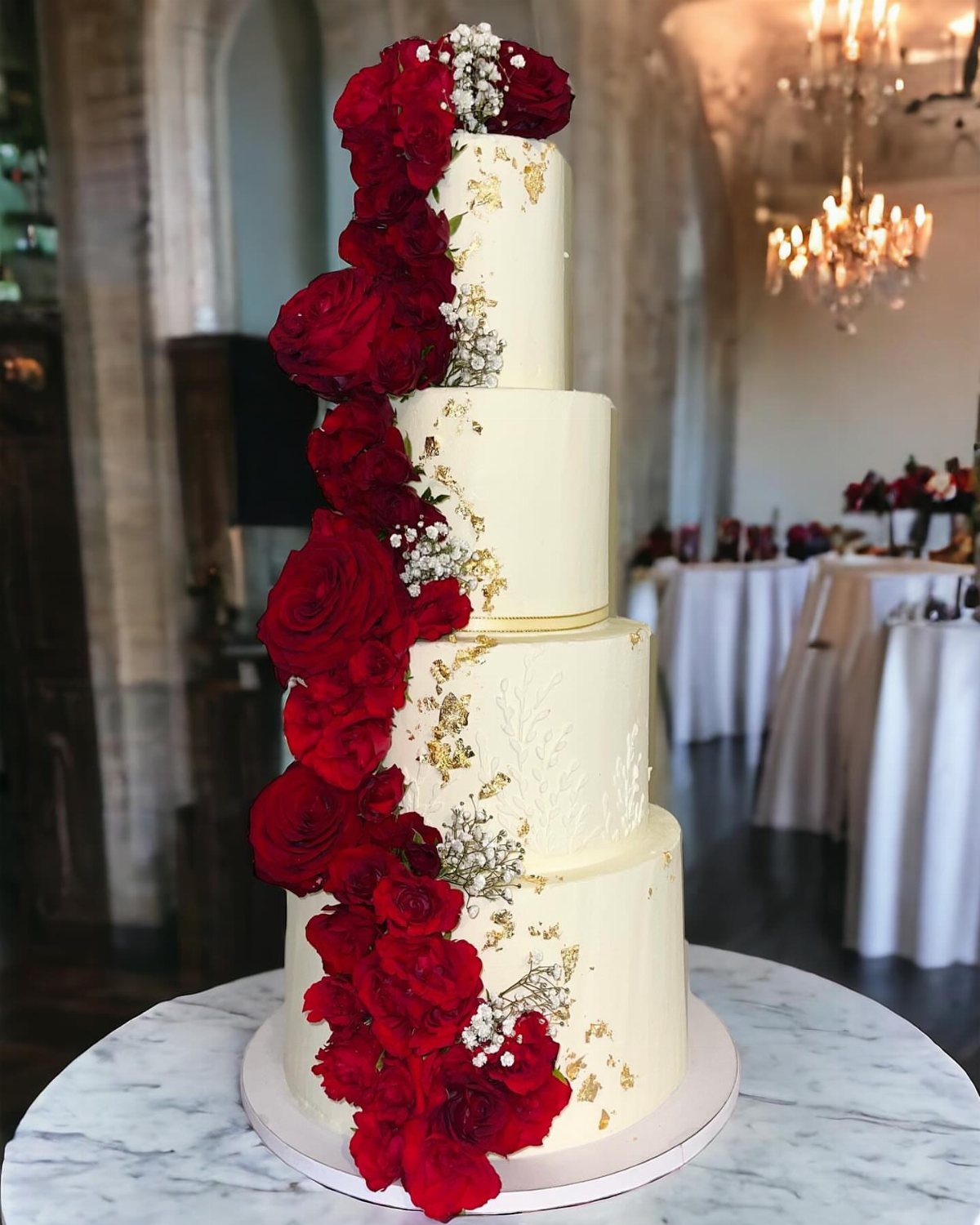 12 Romance Wedding Theme Ideas That Actually Feel Personal 8 Four-tier white wedding cake decorated with gold leaf and cascading red roses on a marble table.