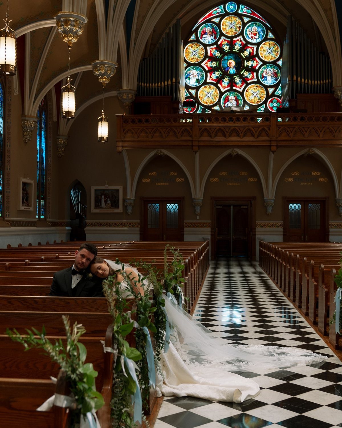 A bride and groom lean against a wooden pew inside a large, ornate cathedral.
