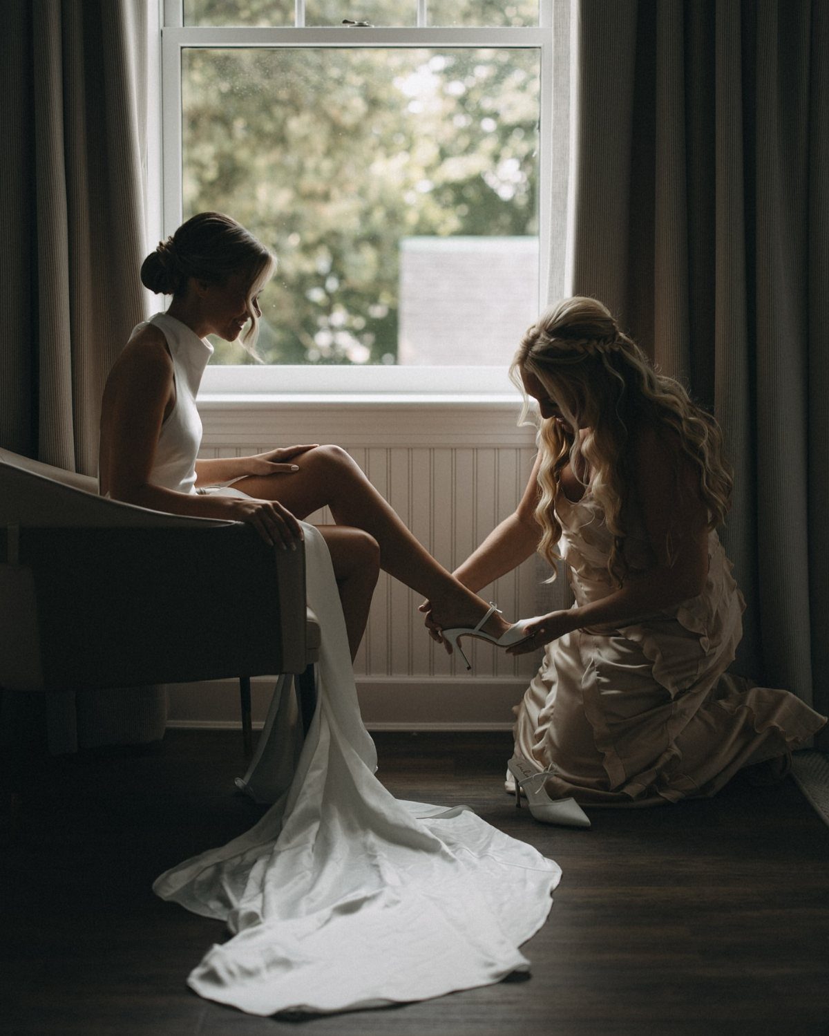 A bride sitting in a chair while a bridesmaid helps her put on a white wedding shoe.
