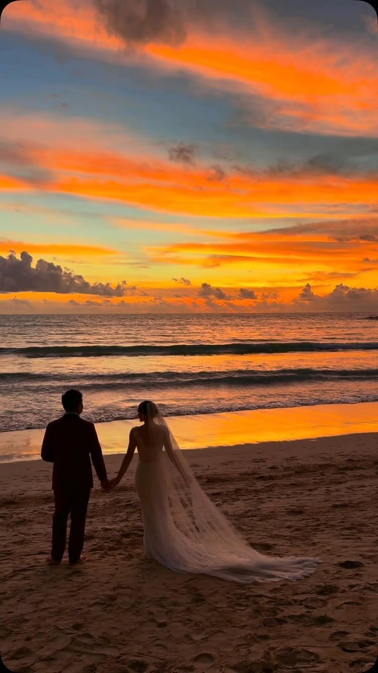 Silhouetted bride and groom holding hands on a beach during a vibrant orange sunset.