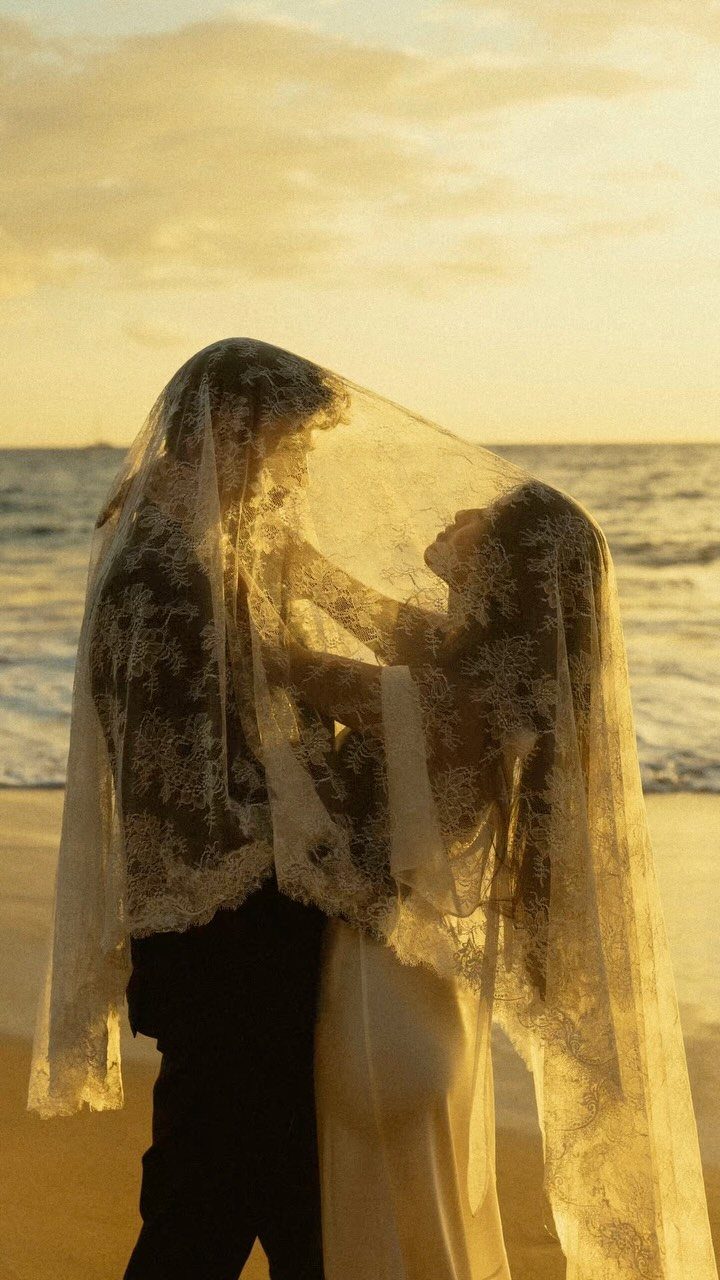 A couple standing on a beach at sunset under a translucent lace bridal veil.
