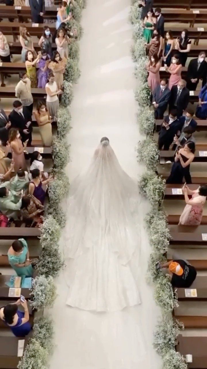 A bride in a white gown walks down a long white aisle runner lined with greenery.