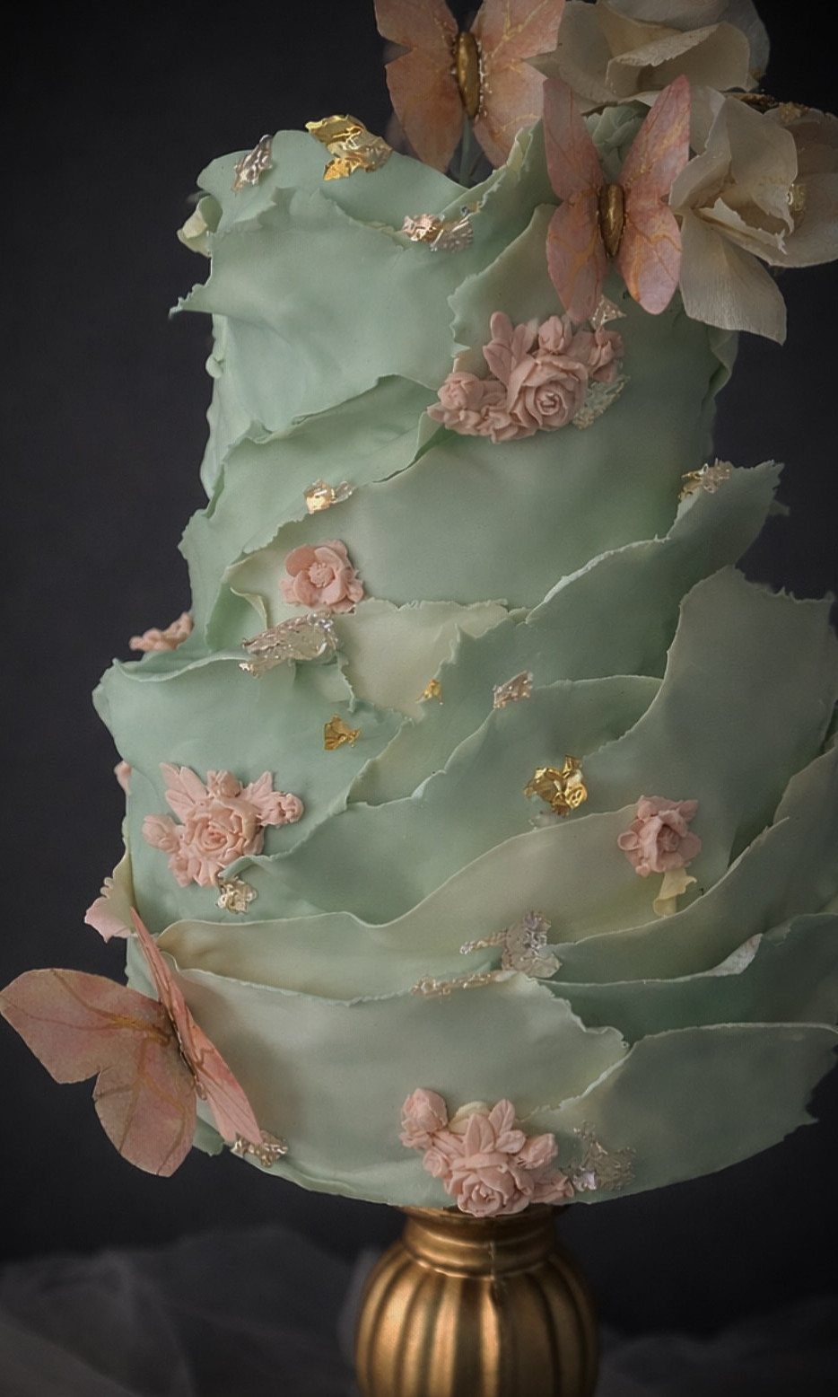 A ruffled pale green cake decorated with pink roses, gold leaf, and peach butterflies.