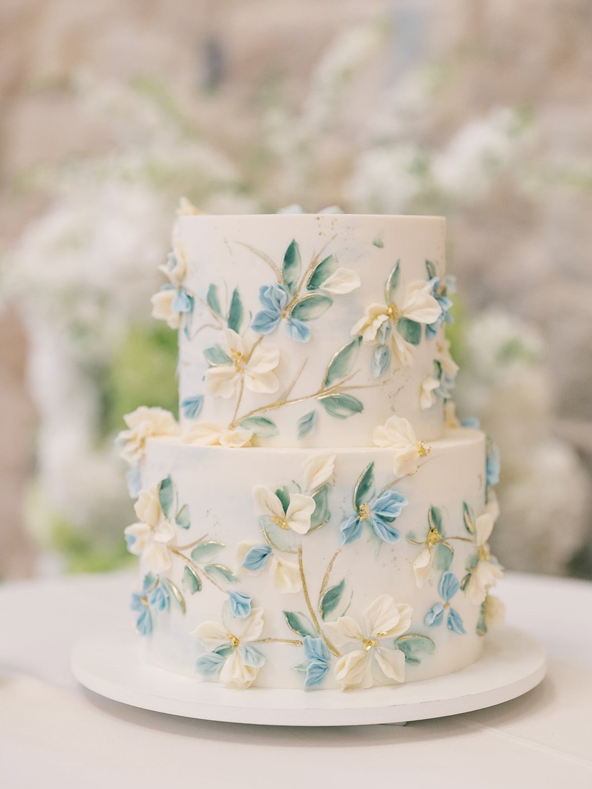 Two-tier white wedding cake decorated with blue and cream floral patterns and gold accents.