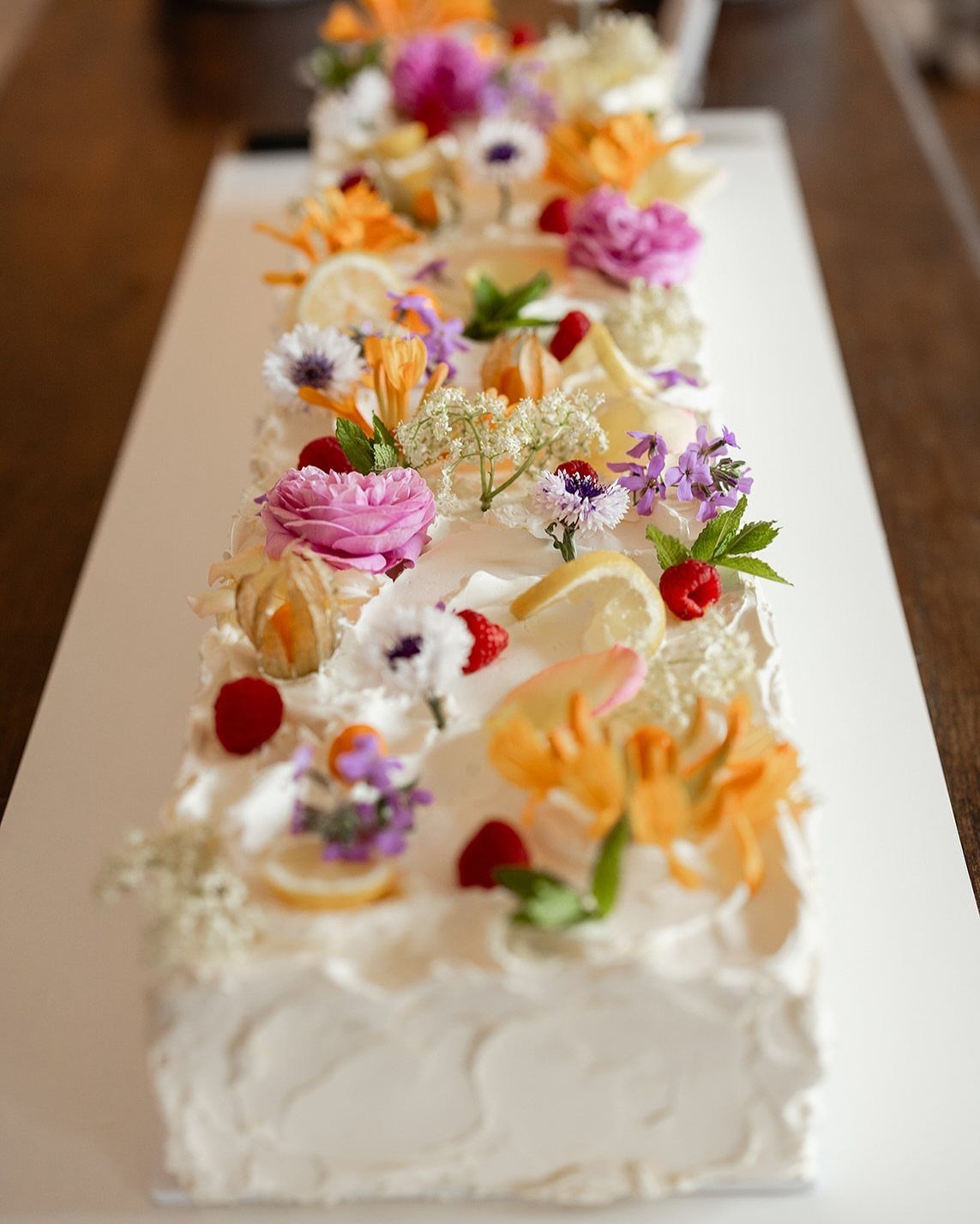 A long rectangular white frosted cake decorated with colorful spring flowers and fresh berries.