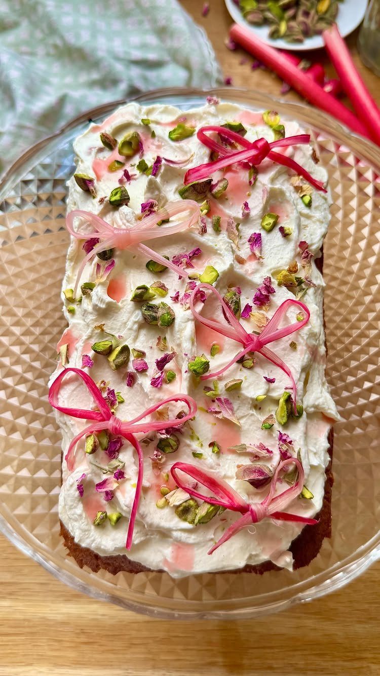 A rectangular loaf cake with white frosting, crushed pistachios, pink petals, and pink ribbon bows.