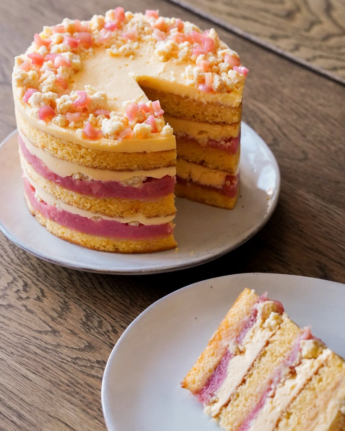 A multi-layered sponge cake with pink rhubarb filling and yellow frosting on a wooden table.