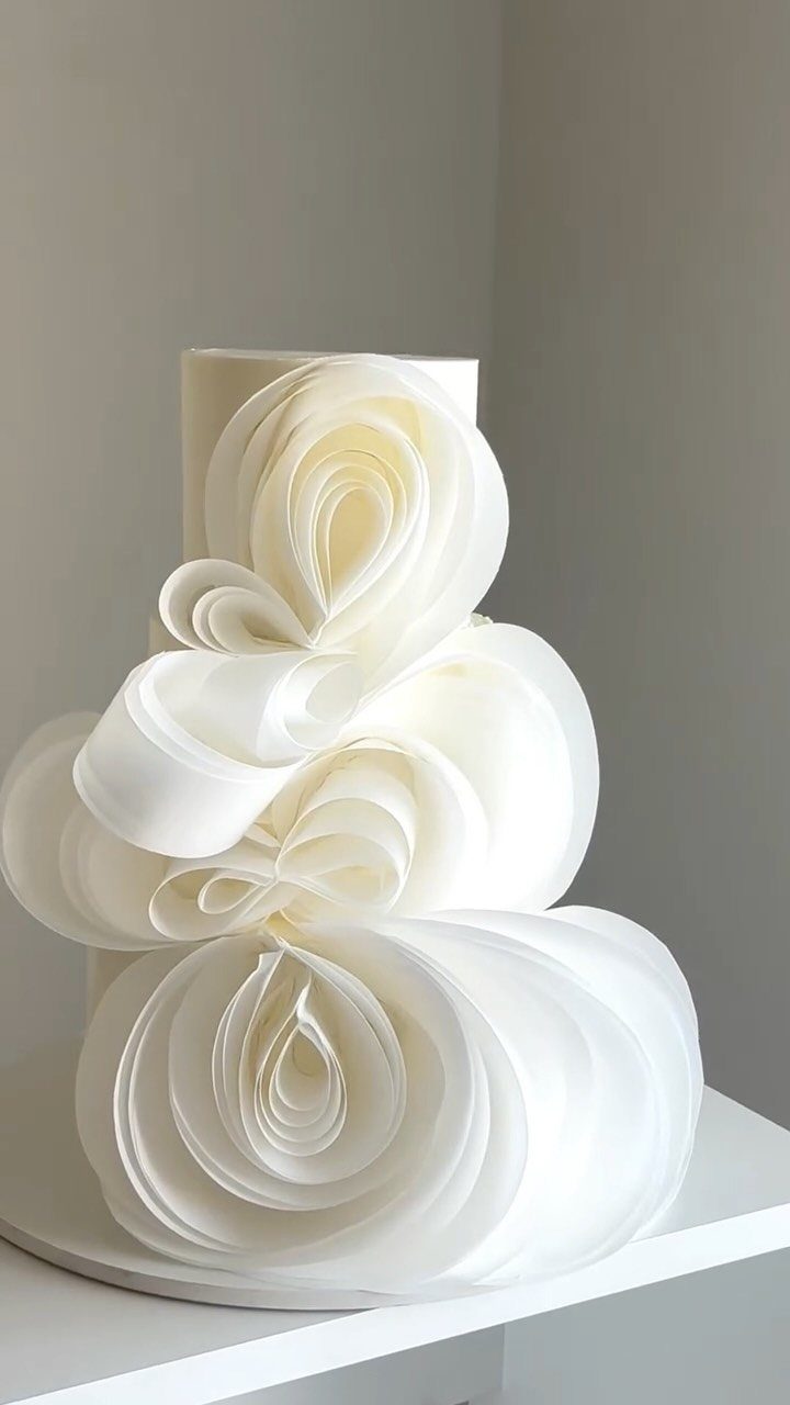 A white tiered cake featuring large, sculptural, flowing frosting swirls resembling organic petals.