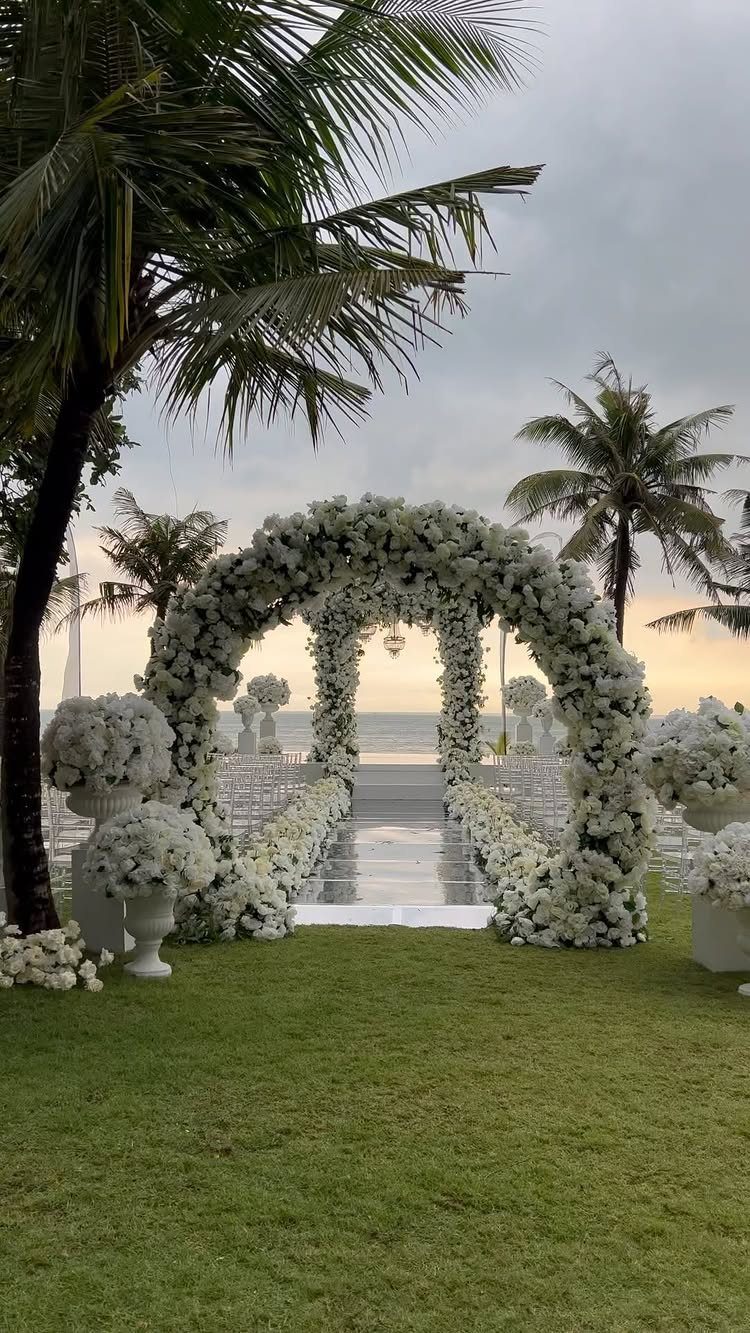 9 Wedding Ceremony Arch Ideas for Indoor and Outdoor Vows 9 A large white floral arch stands on a grassy lawn facing the ocean at sunset.