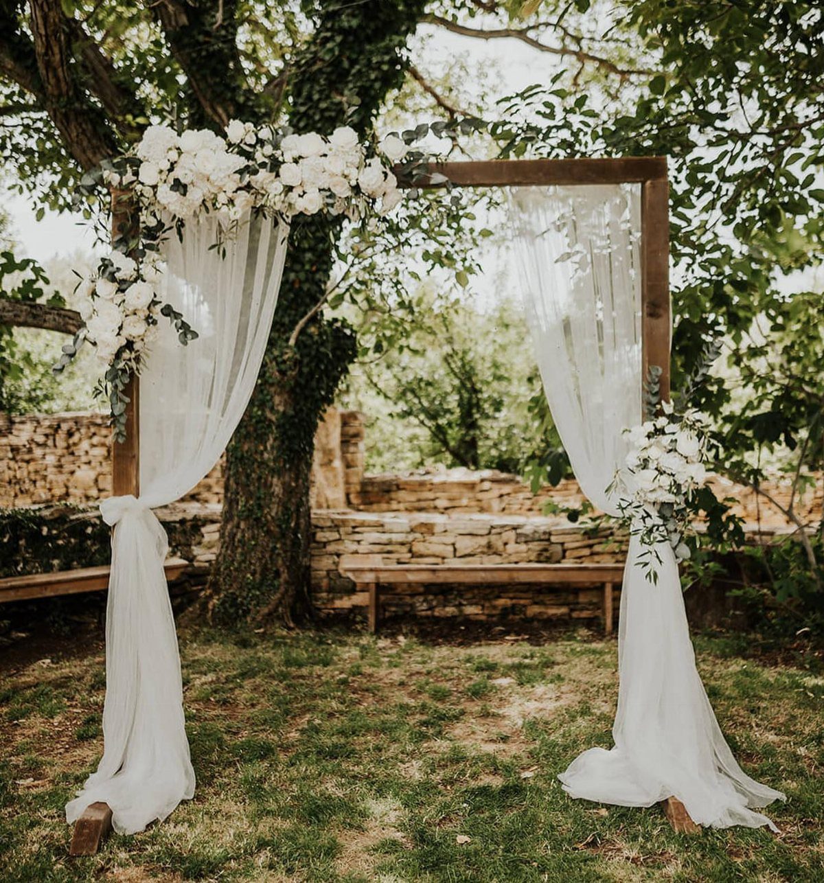 9 Wedding Ceremony Arch Ideas for Indoor and Outdoor Vows 2 A wooden wedding arch decorated with white flowers and draped with sheer white fabric outdoors.
