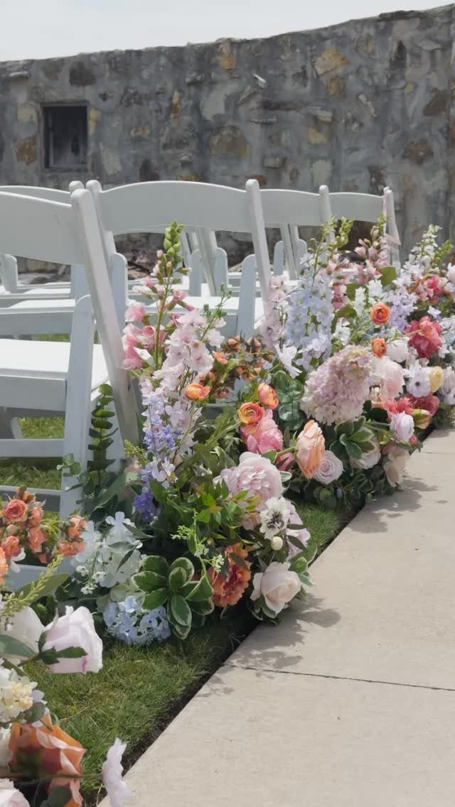 Colorful floral arrangements line a walkway next to white folding chairs outdoors.