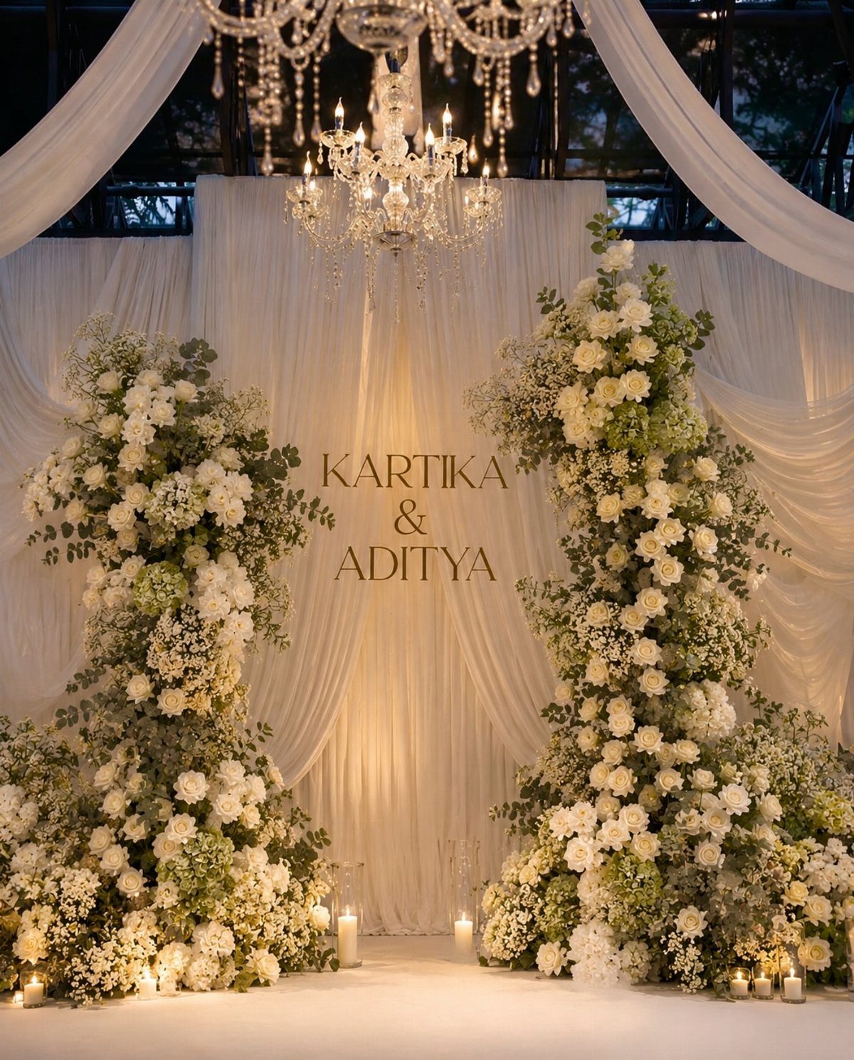 Large white floral arrangements standing against draped white fabric with a crystal chandelier above.