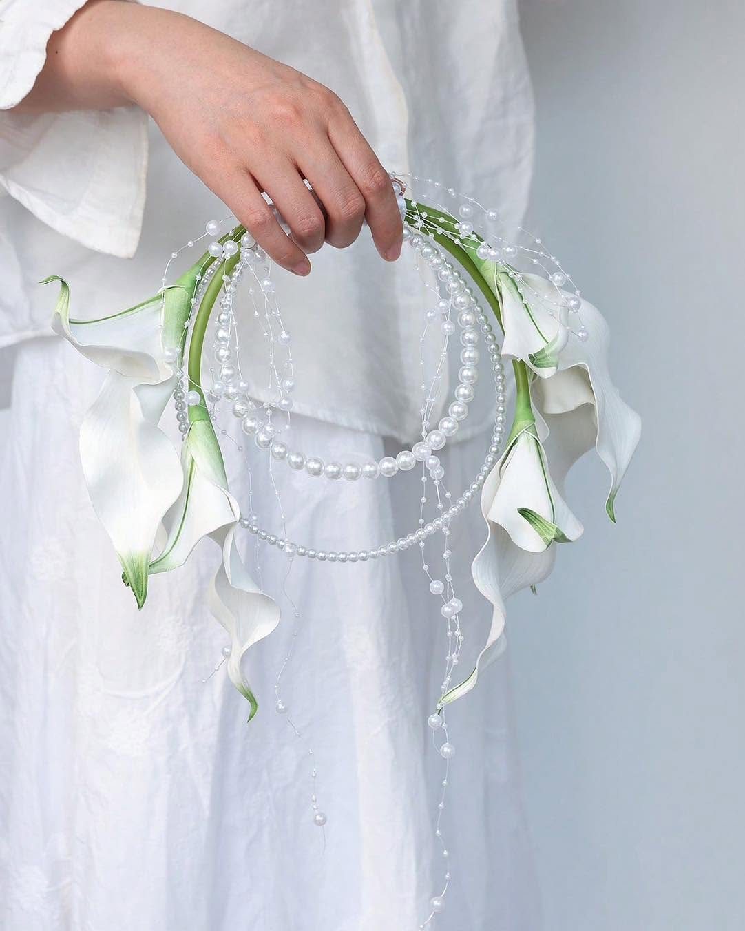 A hand holds a circular bridal bouquet made of white calla lilies and hanging pearls.