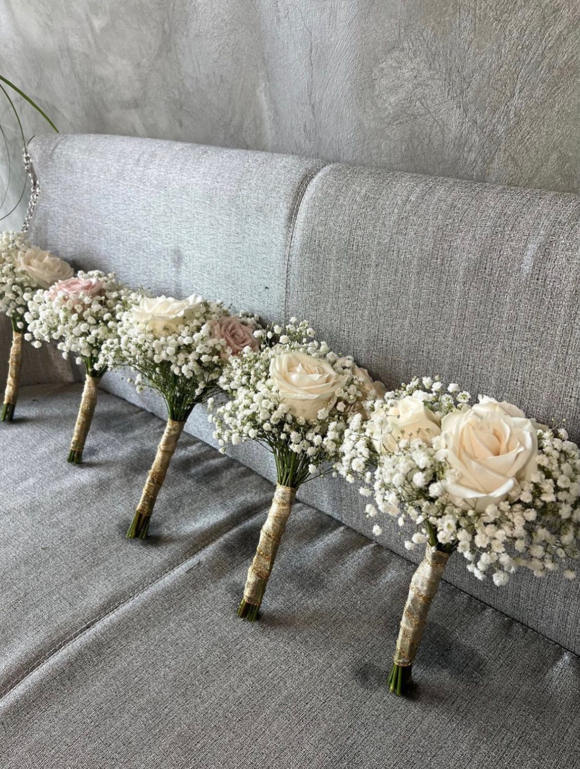 Five small bouquets of white roses and baby's breath with gold wrapped stems on a grey sofa.