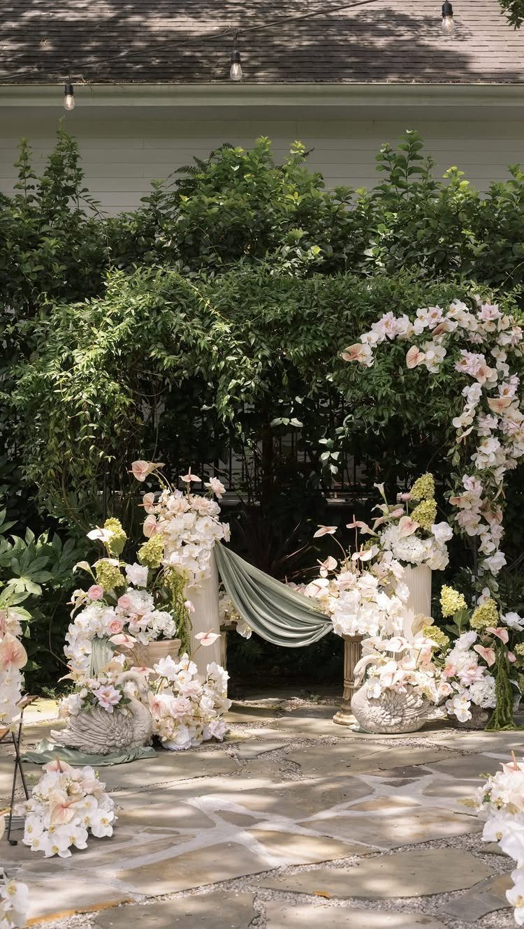White and pink floral arrangements with greenery and draped fabric on a stone patio.