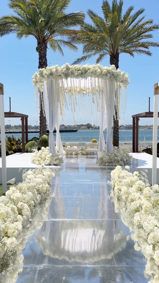 White floral arrangements line a reflective wedding aisle leading to a floral chuppah by the ocean.