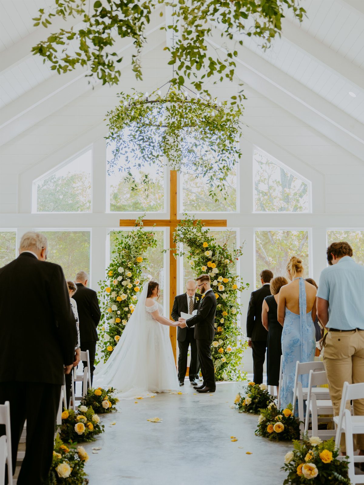 A bride and groom exchange vows before a floral altar and wooden cross in a chapel.