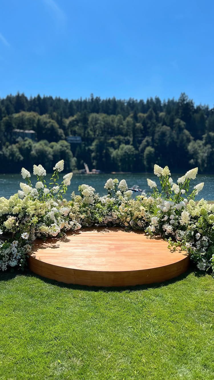 White floral arrangements surrounding a circular wooden platform on a green lawn by a lake.