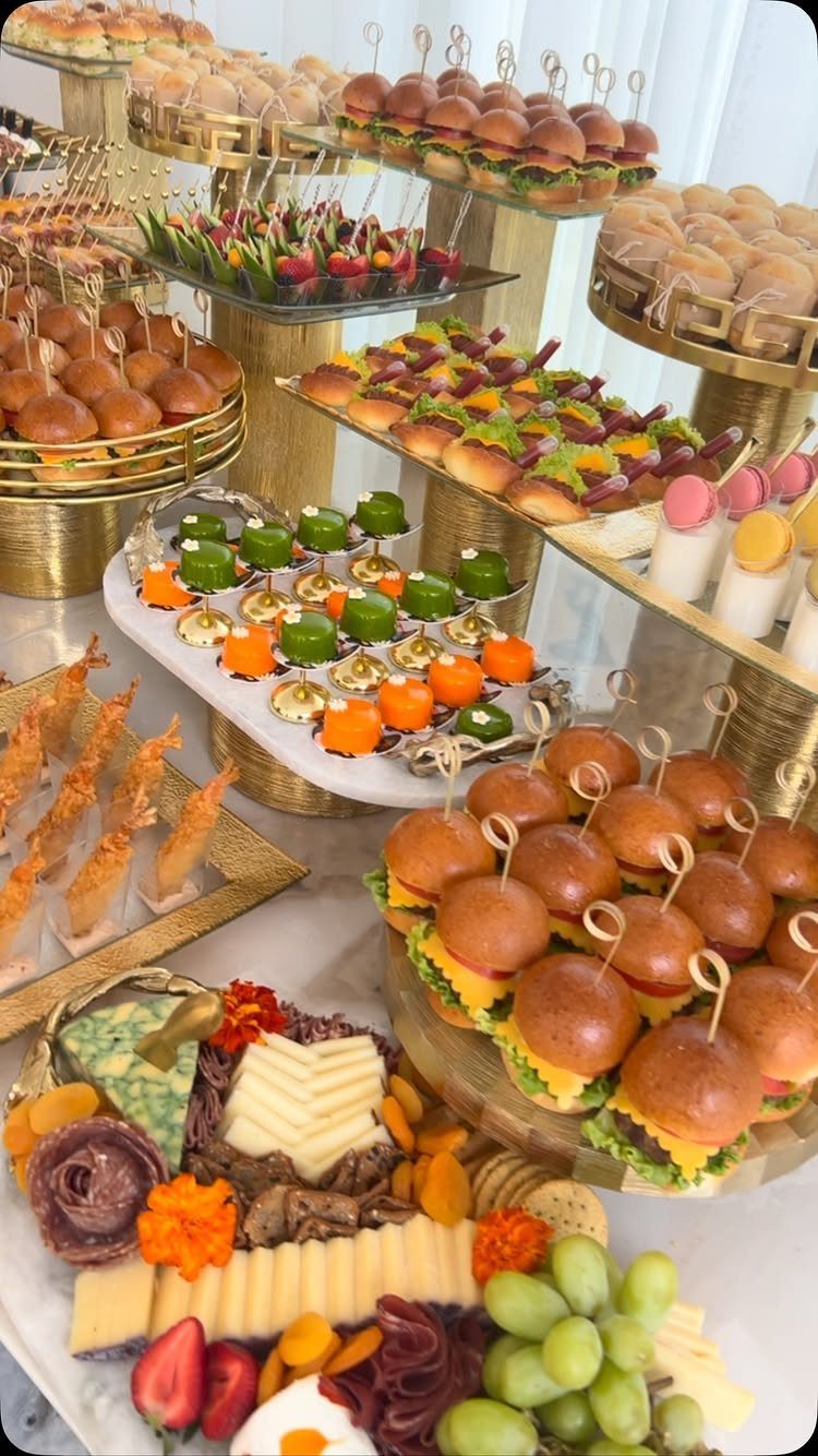 12 Wedding Events Ideas for Your Reception, Ceremony & More 2 A diverse buffet spread with sliders, mini sandwiches, fruit skewers, and a charcuterie board on gold stands.