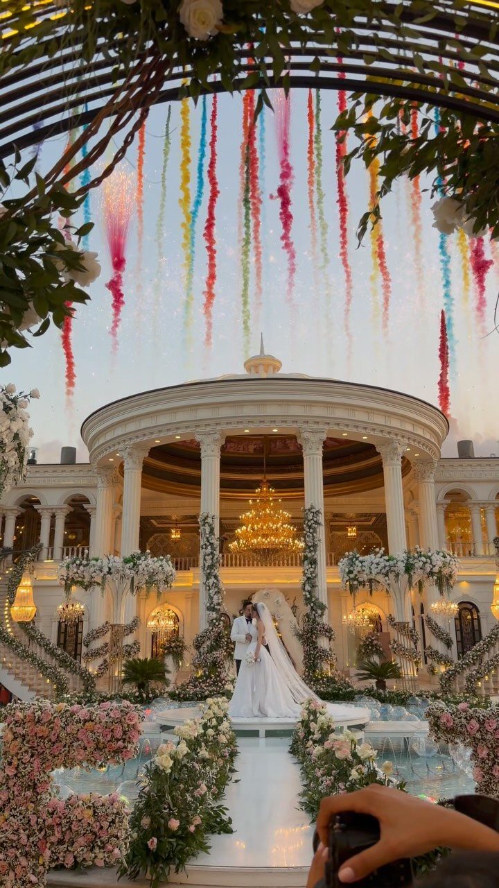 12 Wedding Events Ideas for Your Reception, Ceremony & More 4 A bride and groom standing before a grand white building with colorful smoke trails in the sky.