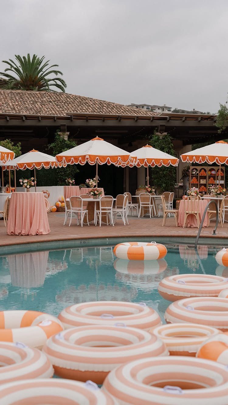 12 Wedding Events Ideas for Your Reception, Ceremony & More 7 Orange and white striped umbrellas and tables set up beside a swimming pool with matching inflatable rings.