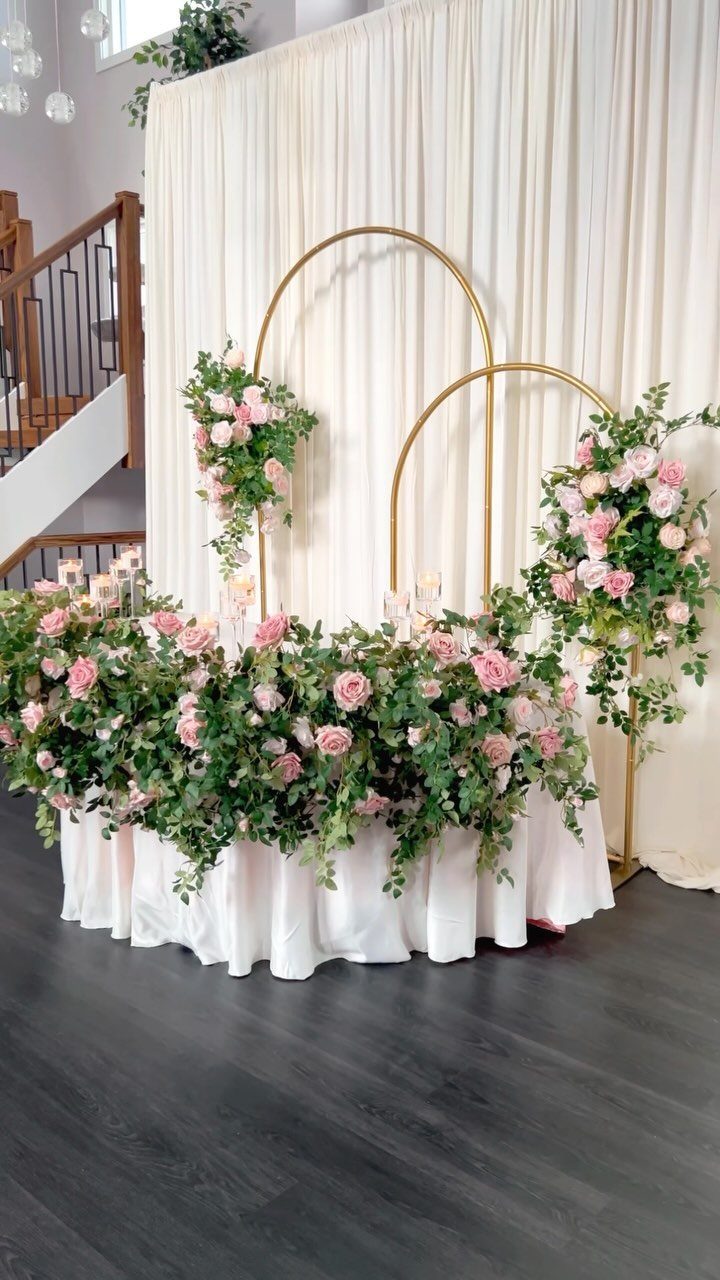 12 Wedding Events Ideas for Your Reception, Ceremony & More 11 A wedding sweetheart table decorated with pink roses, greenery, and gold circular arches against white drapes.