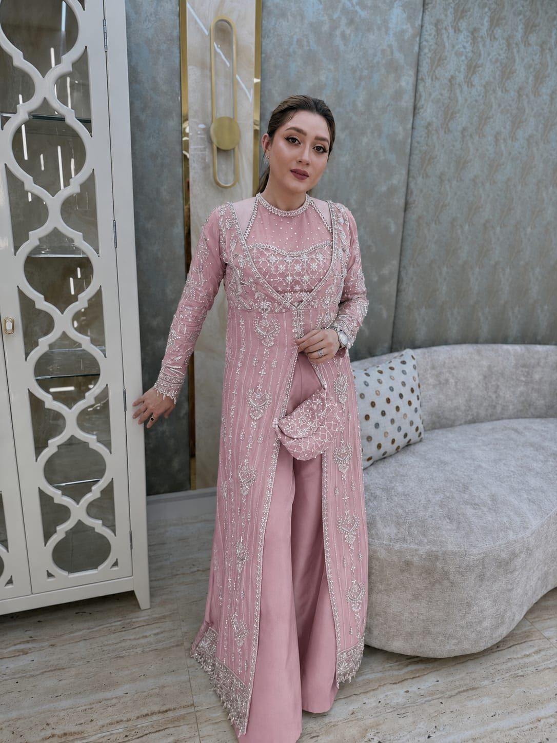 Woman in an embellished dusty pink long tunic and wide-leg trousers standing in a modern room.