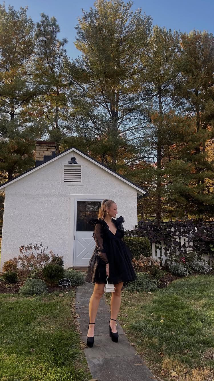 Woman in a voluminous black dress and heels standing on a path in front of a white house.