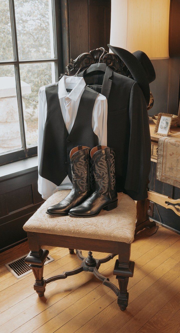 13 Wedding Ideas for Country Ceremonies That Feel Lived-In 8 Black leather cowboy boots resting on a cushioned stool next to formal western wedding attire.