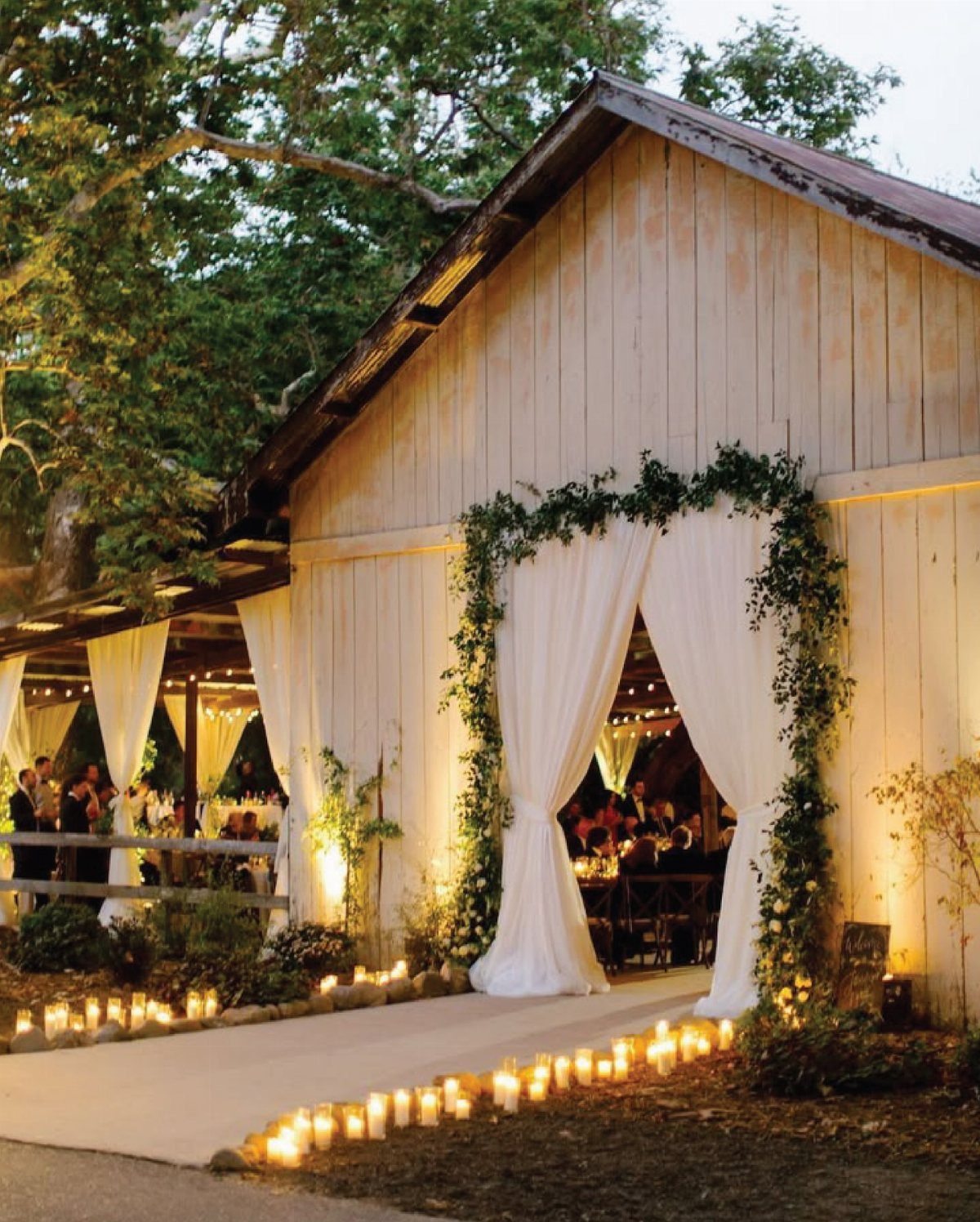 13 Wedding Ideas for Country Ceremonies That Feel Lived-In 9 A rustic wooden barn decorated with white drapes, greenery, and glowing candles along the entrance path.
